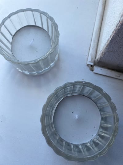 The image shows two small, clear glass votive candle holders on a white surface, likely a windowsill. Each holder contains a white tea light candle. The candle on the left is positioned slightly further back and towards the top of the frame, while the candle on the right is closer and towards the bottom. The glass of the holders is ribbed vertically and has a scalloped edge. In the upper right corner, a portion of a white, textured object is visible, which appears to be a window frame or sill. The overall lighting suggests it is daytime. There are no people or animals in the image. The scene is static and appears to be an indoor setting. The location is not discernible from the image itself, but it has been provided as Osek, Czechia.