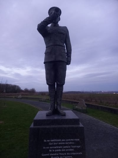 A bronze statue of a soldier stands at attention, saluting with his right hand near his head. He wears a uniform, including a hat, jacket, and boots. The statue is mounted on a stone base. A granite plaque behind the statue bears an inscription in French: "Ils ne vieilliront pas comme nous Qui leur avons survécu: Ils ne connaîtront jamais l'outrage Ni le poids des années. Quand viendra l'heure du crépuscule Et celle de l'aurore," which translates to "They shall not grow old as we that are left grow old: Age shall not weary them, nor the years condemn. At the going down of the sun and in the morning, We will remember them." Grass and a bench are visible in the background, with a skyline in the distance under a cloudy sky.