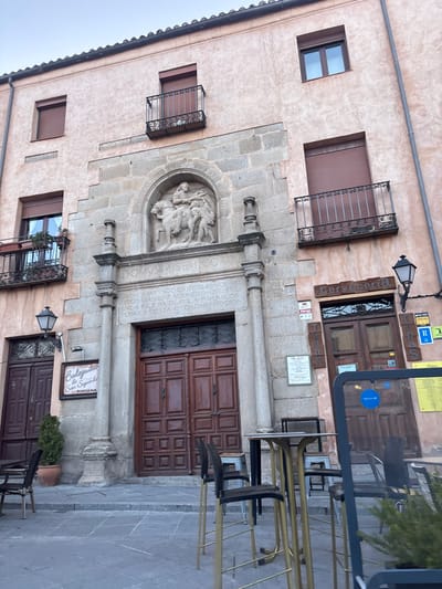The image displays the facade of a multi-story historic building in Ávila, Spain, centered around a stone entrance portal. The building features a light pinkish-ochre stucco exterior contrasted with grey stone blocks forming the central section and quoins. The central portal consists of large, dark brown, double-leaf wooden doors with recessed panels, flanked by two fluted stone columns. Above the doors, an inscribed stone lintel reads "DOMVS MISERICORDIAE" and additional Spanish text, indicating the founding of a church in Ávila for the city's poor. Directly above this inscription, within a rounded arch, is a stone relief sculpture depicting a figure on horseback engaging with another kneeling figure.
The building's upper levels feature several rectangular windows, most equipped with closed brown louvered shutters, and wrought-iron balconies. On the ground level, to the left of the central portal, is an entrance for "Bodeguita San Segundo," marked by a dark wooden door and a wall-mounted streetlight. A slender potted plant stands near this entrance. To the right of the central portal, another entrance for a "Cervecería" (Brewery/Pub) is visible, with a dark wooden door and a corresponding streetlight, featuring vertical "Tapas" and "Copas" signs. The foreground reveals a paved outdoor area with several high-top tables, characterized by golden bases and dark seating, suggesting an outdoor dining or drinking space. A translucent partition with a blue circular logo is partially visible in the lower right. No people are present in the scene. The sky above is pale and uniformly lit.