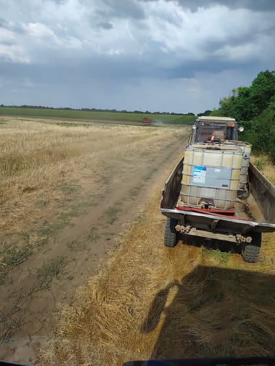 In a rural setting, likely in Alexandru Ioan Cuza, Moldova, a trailer is being pulled by a tractor through a field of dry, yellow stubble. The trailer carries two large white plastic containers filled with liquid, likely agricultural chemicals, as indicated by a label on one of the containers. The trailer is attached to a tractor, the front of which is visible in the foreground. In the background, another red tractor is working in a green field under a cloudy sky. The overall environment appears to be agricultural, possibly during harvest season. The scene is captured from a perspective slightly above and behind the trailer.