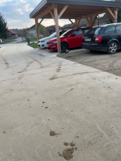 The scene depicts a residential area in Krško, Slovenia, under an overcast daylight sky. The foreground features a light-colored concrete surface marked with numerous tire tracks and localized deposits of mud, notably concentrated in the lower right quadrant. A wooden carport, supported by light-colored timber posts and featuring a dark roof, occupies the midground. Three passenger vehicles are parked under or immediately adjacent to the carport, all oriented away from the camera. From left to right, these include a white sedan, a red hatchback with dark, multi-spoke wheels, and a black station wagon. A small, dark object, possibly a car jack or tool, is positioned on the ground near the front left wheel of the red vehicle. Between the foreground and the parked cars is a strip of unpaved earth. The background reveals a paved road curving into a residential setting, with multiple houses and trees visible under a partially cloudy sky. No persons are present in the image.