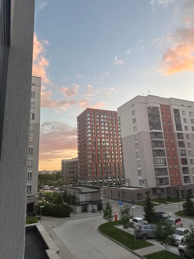 This media file depicts a view of modern residential buildings in Koltsovo, Russia. The image is oriented vertically, showing multiple apartment blocks of varying heights and architectural styles. The buildings are predominantly light-colored with darker brick accents and numerous windows. In the foreground, a textured concrete ledge is visible, suggesting the image was taken from a balcony or window.
The scene includes a paved area with parked cars, lined with small trees and green lawns. A blue parking sign with a white "P" is visible. The sky is a soft blue, with scattered clouds tinged with orange and pink, indicating either sunrise or sunset. The lighting suggests it is late afternoon or early evening.
A few indistinct figures can be seen walking on the paved area between the buildings and cars. The overall atmosphere is calm and residential. The location is explicitly stated as Koltsovo, Russia. There are no discernible emotions conveyed, and the image focuses on the urban landscape. No visible text other than the parking sign.