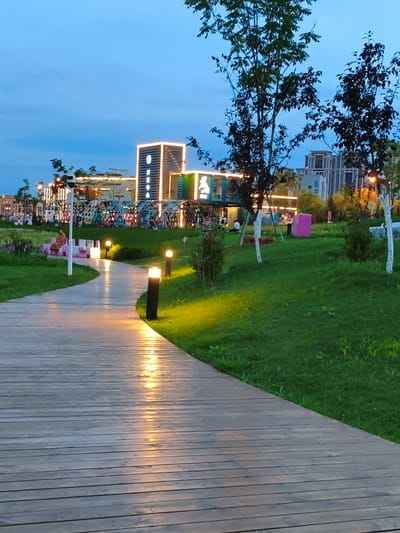 The image depicts an outdoor public space in Jiguan District, China, at dusk. A wooden walkway curves through a grassy park area, illuminated by warm-toned pathway lights. In the background, a modern building with illuminated signage stands prominently, surrounded by trees and other structures. Several people are visible in the distance, some sitting on benches and others standing. The sky is a deep blue, indicating the transition from day to night.