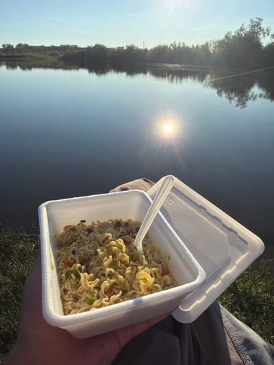 In Taskino, Russia, a person is holding a white styrofoam container of instant noodles by a calm body of water. The sun is low in the sky, casting a warm glow and creating a reflection on the water's surface. Trees and vegetation line the opposite bank of the water, and the sky is clear and blue. The noodles appear to be prepared with vegetables and possibly some meat. A plastic fork is in the container. The person is wearing light-colored pants. The scene suggests a peaceful moment of enjoying a meal outdoors by the water.