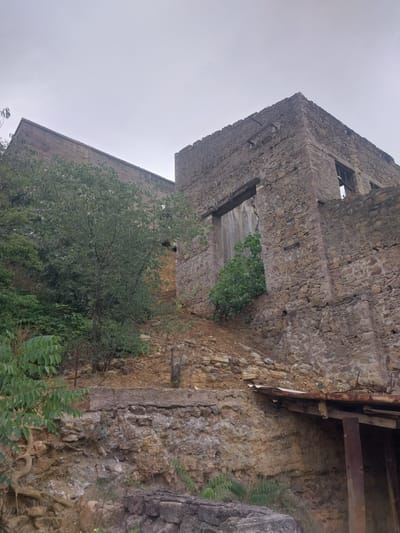 The image depicts the exterior of an abandoned stone building in Kapan, Armenia. The structure appears old and weathered, with rough-hewn stone walls and open, broken windows. Overgrown vegetation, including trees and vines, encroaches on the building, suggesting a long period of disuse. One section of the building shows exposed wooden beams and rusting metal supports, hinting at past construction or decay. The ground is rocky and uneven, with loose stones and soil visible. The sky is overcast and a pale gray, indicating a cloudy or foggy day. The overall mood is one of desolation and decay, with nature reclaiming the man-made structure. There is no visible text in the image.