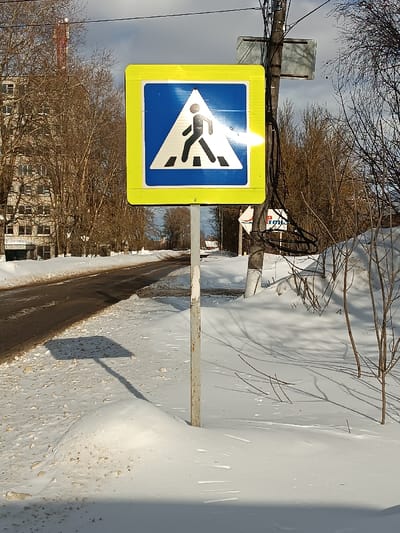 The image displays a winter street scene in Furmanov, Russia, illuminated by partial sunlight. A square pedestrian crossing sign, featuring a yellow border, a blue inner square, a white triangle, and a black walking pedestrian symbol, is positioned prominently on a metallic pole in the foreground. A bright glare from direct sunlight is visible across the center of the sign. The pole is embedded in a snow-covered bank to the right of an asphalt road. The road, which shows dark pavement with brown slush and dirt along its edges, curves to the left into the distance.
To the right of the pedestrian sign, a dark utility pole supports multiple cables. Attached to this utility pole is a rectangular, reflective sign above, and a smaller white sign with red Cyrillic text, partially legible as "тройка" (troika), below. The ground throughout the scene is covered in a substantial layer of snow. Bare deciduous trees line both sides of the road and form a dense background. On the far left, beyond the trees, a multi-story, light-colored residential building is visible, with a red and white striped antenna or tower extending above it. The sky is partly cloudy, and the pedestrian sign casts a distinct square shadow on the snow to its left. No individuals are present in the visible area.