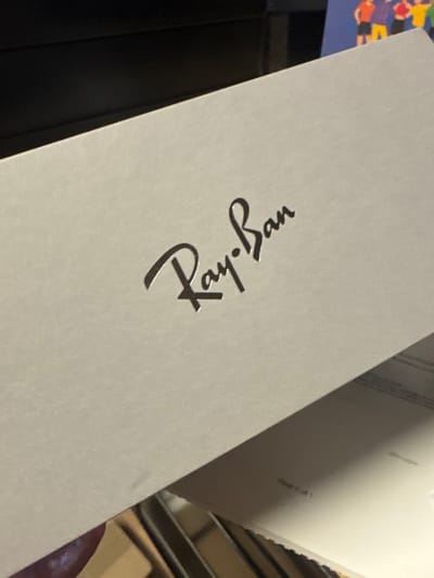 A close-up shows a white box with the Ray-Ban logo printed in black. The logo is diagonally oriented, with "Ray" positioned below and "Ban" above. The surface of the box appears smooth and matte. A stack of white papers, potentially documents or manuals, are partially visible behind the box. A hint of a colorful object, perhaps a book or artwork, can be seen in the upper left corner.