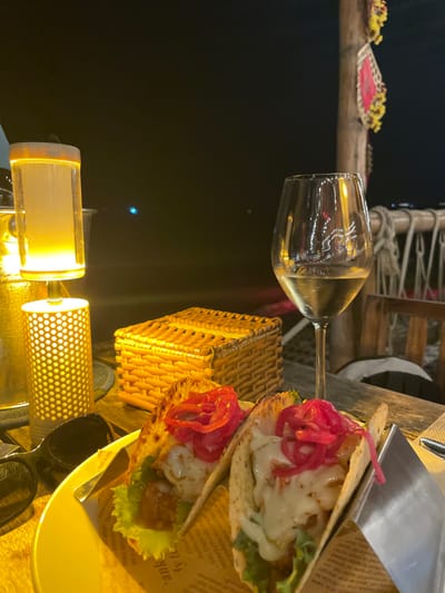 The image depicts a nighttime outdoor dining scene at a restaurant in Thành phố Phan Thiết, Vietnam. In the foreground, two soft-shell tacos are presented on a circular yellow plate lined with paper. Each taco contains green lettuce, a fried protein, a white sauce or melted cheese, and vivid pink pickled red onions. To the right of the tacos, a clear stemmed wine glass holds white wine, reflecting ambient lights.
To the left of the plate, a decorative table lamp provides warm illumination, featuring a cylindrical perforated base and a taller cylindrical shade. Adjacent to the lamp is a metallic, possibly gold-toned, cylindrical container. A pair of black-framed sunglasses rests on the table near the lamp. Further back on the table, between the lamp and the wine glass, is a small, rectangular, woven basket-like box.
The objects are positioned on a rustic wooden table surface. The background is a dark expanse, consistent with a body of water at night, dotted with scattered, distant lights. To the far right, a wooden post adorned with blurred red and yellow decorative elements is visible, likely part of the restaurant's structure or decor, with a rope railing faintly discernible below it. No individuals are visible in the scene.
