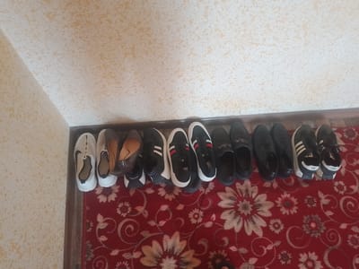 In Vahdat, Tajikistan, a hallway features a shoe rack overflowing with various footwear. On the left, a pair of white Nike sneakers sits beside brown loafers and a pair of dark slip-on shoes. The right side of the rack displays a collection of black shoes, including sneakers with white stripes and loafers. The shoes are arranged haphazardly on the rack, with some pairs overlapping. The hallway is decorated with a red carpet featuring a floral pattern. The walls are a light yellow color with a textured finish, and a wooden baseboard runs along the bottom of the walls. The lighting in the hallway is dim, casting shadows on the floor and walls. The scene suggests a functional space where shoes are stored, with a touch of personal style evident in the variety of footwear.