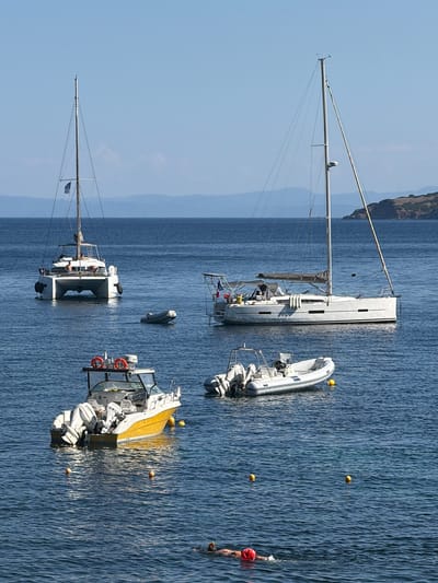 This media file captures a serene scene at sea, likely off the coast of Skiathos, Greece. The image features several boats anchored in clear, deep blue water. The main subjects are a large white sailing yacht with its mast prominently displayed, and a smaller, white inflatable boat with outboard motors. Another smaller white boat with a cabin and life rings is also visible. In the distance, a hilly, treed shoreline can be seen under a clear, bright blue sky, suggesting a sunny day with good weather.
A person in the water, wearing a red cap and possibly swimming or treading water, is a notable detail. Several yellow buoys are scattered across the water, indicating a marked area or mooring points. The overall impression is one of a peaceful anchorage, with the boats resting on calm waters. The lighting and clear visibility suggest the image was taken during daylight hours, likely mid-morning or afternoon. There is no visible text in the image. The scene evokes a sense of tranquility and leisure associated with a day at sea.