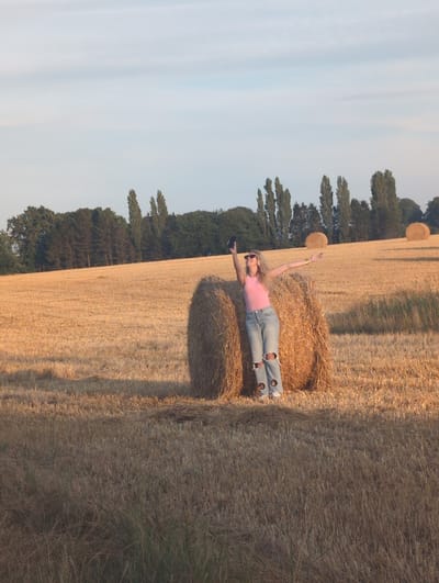 In Alvimare, France, a young woman with blonde hair stands in a field of dry, golden stubble under a pale blue sky with scattered white clouds. She is leaning against a large, round bale of hay, her left arm extended upwards, holding a black smartphone as if taking a selfie. Her right arm is also raised, open to the side. She wears a bright pink sleeveless top and distressed light blue jeans with large cutouts on the knees. The field is dotted with several other hay bales, and a line of trees forms the background, with some taller, slender trees interspersed with denser foliage. The lighting suggests it is late afternoon or early evening, casting long shadows and illuminating the scene with a warm, golden hue.