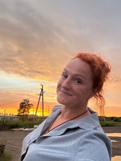 This is a vertical, close-up selfie of a woman with vibrant red hair, taken outdoors during sunset. The woman is facing the camera, her head tilted slightly to the left, with a subtle smile and her gaze directed towards the lens. Her red hair is styled in loose, wispy curls that frame her face. She is wearing a light blue, collared shirt with the top button undone, revealing a dark string necklace.
The background of the image is dominated by a dramatic sunset sky filled with clouds painted in hues of orange, yellow, and soft gray. The light from the setting sun creates a warm glow. In the mid-ground, silhouetted against the sunset, are utility poles with power lines stretching across the frame, and a solitary tree with a sparse canopy. Beyond the tree, there are hints of buildings and greenery, suggesting a rural or semi-rural setting. The ground in the foreground appears to be a dirt or gravel surface, possibly with some puddles reflecting the sky.
The time of day is clearly sunset, evidenced by the warm, low-angled light and the colorful sky. The weather appears calm, with the clouds indicating a beautiful evening. The presence of utility poles and the general landscape suggest a location in Russia, specifically Sovkhoznyi, as indicated by the provided context. The woman's expression conveys a sense of calm contentment. There is no visible text in the image.