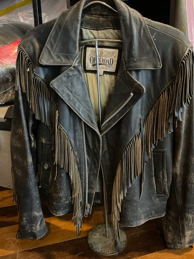 A dark, heavily worn leather jacket, possibly black or very dark brown, hangs on a tarnished metal display stand. The jacket is styled as a biker jacket with wide, notched lapels and an asymmetrical silver-toned zipper closure extending diagonally from the left side. Extensive leather fringes, which appear lighter brown or discolored from wear, are attached along the shoulder seams, across the chest, and along the sleeves. The leather surface of the jacket exhibits numerous scuffs, scratches, and patches of lighter-colored dust or debris, indicating significant age and use. The sleeves, lower front, and collar show particular signs of wear and discoloration. An internal label, light-colored with dark text, is visible at the neck area, displaying "OPEN ROAD" with an eagle graphic above it and two stars, along with smaller text that appears to indicate "MADE IN" and "SIZE 40." Below the jacket, a wooden floor with visible grain and planks is present. In the background, out-of-focus elements include a clear plastic bag containing a red object and indistinct dark structures. No people are visible, and no actions or interactions are depicted. The scene is believed to be located in Ban Yid, Thailand, though specific identifying geographical features are not discernible within the image.