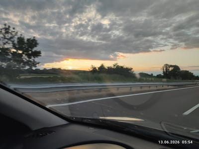 The image captures a view from inside a vehicle, looking out through the windshield at a highway scene at sunrise. The sky is filled with dramatic, grey clouds, with a sliver of bright orange and yellow sunlight breaking through near the horizon on the left side of the frame. The road ahead is paved and appears to be a multi-lane highway, with a white line marking the lanes. A metal guardrail separates the highway from a grassy verge and a line of trees in the distance. The interior of the car, including the dashboard and a portion of the windshield's reflection, is visible in the foreground. The timestamp in the lower right corner indicates the date and time as 13.06.2026 05:54. There are no people or distinct actions depicted. The scene suggests a journey on a highway in or near Shtarkovo, Bulgaria, during early morning hours as the sun rises.