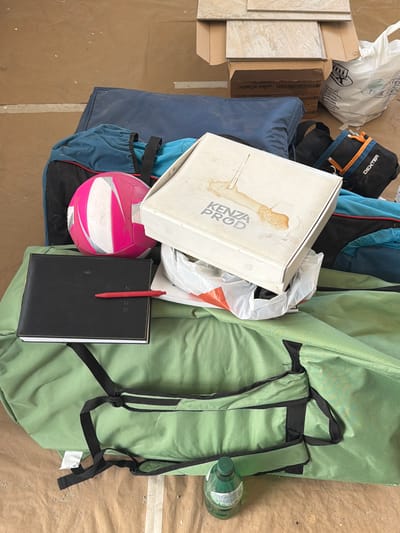 The image shows a jumble of items on a brown, textured surface, possibly a floor or ground. The scene is cluttered and appears to be an outdoor or storage area.
Prominent objects include a white cardboard box with "KENZA PROD" printed on it, a pink and white volleyball, and a black notebook with a red pen inserted. There are also several bags: a blue bag, a black bag with orange and blue accents and "DEXTER" text, and a large green bag with black straps, resembling a backpack or duffel bag. A plastic bottle with a green cap and label is visible near the green bag. Additionally, there are some light-colored tiles and cardboard boxes in the background.
The items are arranged in a disorganized manner, suggesting they might be packed for transport, in storage, or have been recently unpacked. There are no people visible in the image. The lighting appears natural, but there are no specific cues about the time of day or weather. The overall impression is one of a collection of personal belongings or equipment. The visible text includes brand names like "DEXTER" and "KENZA PROD", and the number "2025" on the notebook. The location context is Gondomar, Portugal.