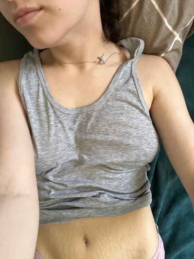 The image shows a close-up, overhead view of a person lying down. The person is wearing a grey tank top and a silver necklace with a small, abstract pendant. The lower abdomen, with visible stretch marks, is exposed. The background consists of a dark green blanket and a brown, diamond-patterned fabric. There are no identifying landmarks or features that would suggest a specific location within Postoyata, Ukraine.