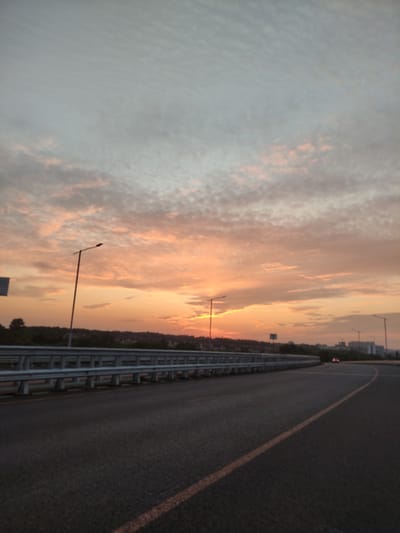 A vertical, low-angle, medium shot captures a road and a scenic sunset over Kraskovo, Russia. The sky is filled with a dramatic display of clouds painted in hues of orange, pink, and grey, indicating the sun is setting or rising. A long stretch of asphalt road dominates the foreground, with a yellow line marking the right lane, curving gently to the right. On the left side of the road, a metal guardrail runs along the edge, separating the road from the elevated ground beyond. A single street lamp stands tall on the left, its dark silhouette stark against the vibrant sky. In the distance, a line of trees forms a dark band along the horizon, interspersed with the faint outlines of buildings and more street lamps. A solitary car with its taillights illuminated is visible on the road in the far distance, adding a sense of movement to the otherwise static scene. The overall atmosphere is peaceful and serene, characteristic of the natural beauty of Kraskovo at dusk or dawn.