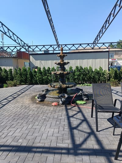 This is a daytime, outdoor, medium shot image taken in the city of Hlobyne, Ukraine, featuring a multi-tiered stone fountain as the central focus. The fountain is situated on a paved patio made of gray rectangular bricks laid in a herringbone pattern. The fountain's tiers are dark brown and appear to be made of weathered stone, with water cascading down them.
Around the fountain, there are some decorative elements, including what appear to be sculpted creatures at its base. A red inflatable ladybug toy and a green mat are present near the fountain's base, along with a pink garden hose.
Behind the fountain, a row of neatly trimmed, dark green evergreen trees forms a natural barrier. Beyond the trees, a light-colored, corrugated metal fence is visible, with some buildings and structures in the background. To the right of the fountain, a black metal-framed lounge chair with a mesh seat is positioned on the patio, casting a long shadow. A small portion of a dark table is also visible in the lower right corner of the frame.
Overhead, a dark metal framework, possibly part of a canopy or pergola structure, extends across the upper portion of the image, creating a crisscross pattern of shadows on the patio. The sky is clear and bright blue. The overall scene suggests an outdoor seating or recreational area.