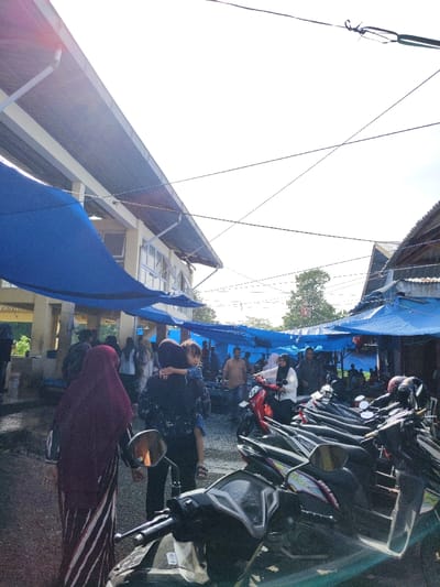 The image depicts an active outdoor market scene in Tuha Gp Gajah, Indonesia, under bright daylight with some lens flare. The perspective is from a slightly elevated position, looking into a crowded market area.
On the left, a multi-story building with a light-colored facade, horizontal windows, and a dark, sloped roof with gutters is visible. Extending from this building and across the scene are multiple blue tarpaulins, providing overhead shade for market activities. On the right, a section of another building with a corrugated metal roof and wooden elements is visible. Numerous utility wires crisscross the bright sky above.
In the immediate foreground, a row of parked motorcycles, predominantly scooters, is aligned on the right, facing deeper into the market. The handlebars and front panels of several scooters are visible. On the left foreground, two women are walking away from the viewer. The woman furthest left wears a dark red hijab and a vertically striped garment. The woman next to her wears a dark, patterned top and carries a young child on her left hip; the child wears a blue top.
The midground and background are populated by a considerable number of people, indicating a busy market environment. Individuals are seen walking, standing, and interacting. A woman in a white hijab and white top is seated on a red scooter in the midground, positioned among other parked motorcycles and market-goers. Additional market stalls, identifiable by more blue tarpaulins and groups of people, extend further into the background. Trees are visible in the distance under the clear sky.