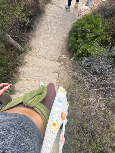 The image depicts a descending outdoor path with steps in Ramatuelle, France. The perspective is first-person, looking down. In the foreground, the upper thighs of an individual are visible, wearing gray athletic shorts on the left and dark brown leggings on the right. A folded olive-green garment rests across the lap, over the brown leggings. A light-colored woven bag, decorated with multiple multicolored fabric flowers (yellow, blue, orange, pink, light green), is positioned on the right thigh. A white shoe is visible at the bottom of the frame, near the right leg. The individual's left hand, featuring long, light-colored manicured fingernails, is visible on the far left.
The path itself is unpaved, composed of compacted dirt and several worn, light-colored steps, showing numerous shoe imprints. Both sides of the path are bordered by dry, scrubby vegetation, including green bushes and clusters of dead branches. On the left side, a weathered wooden post and loose wire fencing are discernible amidst the brush. Further up the path, in the background, two individuals are partially visible, walking away from the viewer. One individual wears dark trousers and light-colored athletic shoes with yellow and black accents. The other individual's lower legs and light-colored shoes are also visible. The environment suggests a coastal or rural hiking trail.