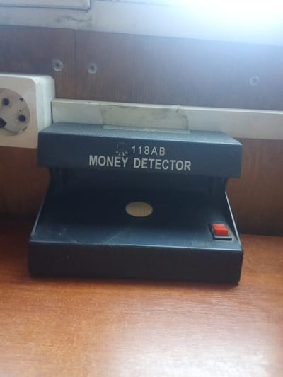 This media file shows a money detector device, likely used for verifying the authenticity of currency. The device is black and has a model number "118AB" printed on its top in white text, along with the words "MONEY DETECTOR." A red power switch is visible on the right side of the device. The detector is placed on a wooden desk, which has a warm, light brown color with a visible wood grain texture.

In the background, a section of a wooden wall is visible, with two screws embedded in it. To the left of the money detector, a white electrical outlet with three sockets is mounted on the wall. The scene appears to be indoors, possibly in an office or a retail setting given the presence of the money detector and electrical outlet. There are no people visible in the image.

The lighting in the image suggests it might be daytime, with a bright light source coming from above and slightly to the right, creating a glare on the upper part of the image and highlighting the texture of the wooden desk. The overall impression is that of a functional setup for currency verification. The provided location context of Alatau, Kazakhstan, suggests where this device might be used.