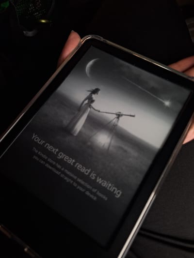A person is holding a Kindle e-reader. The screen displays a black and white image of a woman with a telescope under a starry sky. A crescent moon is also visible in the sky. Text on the screen reads, "Your next great read is waiting" and further describes the Kindle store having a massive selection of books for download. No element of the image indicates it being located in Tlik, Armenia. The background surrounding the Kindle is dark and out of focus, appearing to be clothing.