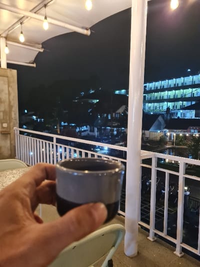 This image captures an elevated nighttime scene in Perbawati, Indonesia. In the immediate foreground, a human right hand holds a two-toned ceramic mug, dark gray at the bottom and lighter gray at the top. A portion of a light-colored, speckled table surface is visible to the left of the hand, and the upper section of a light-colored chair back is seen below. The midground features a white metal railing with vertical balusters, running horizontally across the frame, and a prominent white cylindrical support pillar positioned centrally. Above, a light-colored canopy structure with several exposed incandescent filament bulbs provides overhead illumination. The background reveals a dark night sky above an urban or semi-urban landscape. A large multi-story building with numerous illuminated windows is visible on the right, suggesting an apartment complex or hotel. Various other distant buildings and lights are scattered throughout the background. Below the railing, a ground-level area is visible with multiple parked motorcycles and other vehicles, indicated by their silhouettes and scattered lights.