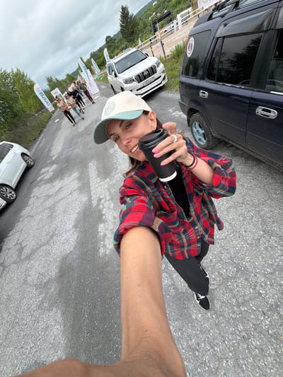 A woman is taking a selfie outdoors while holding a black coffee cup. She is wearing a red and blue plaid shirt, black pants, and a beige baseball cap. She has a white ring on her left ring finger and a red bracelet on her right wrist. In the background, a group of people are walking on a road lined with flags. Several cars are parked on the side of the road, including a white SUV and a dark-colored SUV. The environment appears to be a rural or mountainous area, with trees and hills visible in the distance. The sky is overcast. The location is identified as Теплоозёрск, Russia.