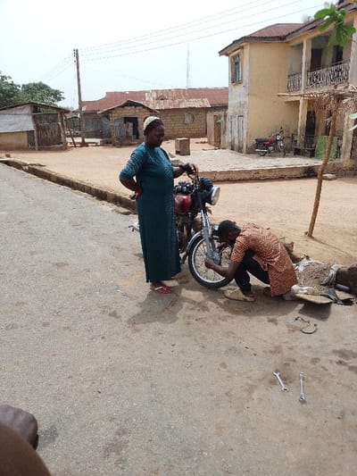 The image depicts an outdoor scene in Igbo Ijaiye, Nigeria, where a motorcycle is undergoing repair on an unpaved shoulder adjacent to a paved road.

**Subjects:**
*   A female adult stands to the left of the motorcycle, facing right, looking towards its front wheel. She wears a dark green, patterned long dress, a patterned head covering, and red flip-flops. Her left hand rests on the motorcycle's handlebar, and her right hand is on her hip.
*   A male adult is crouched on the dirt ground to the right of the motorcycle, engaged in repairing or inspecting the front wheel assembly. He wears a patterned orange and black long-sleeved shirt and dark trousers. His hands are actively manipulating the motorcycle's front wheel area.

**Objects:**
*   A red and silver motorcycle is the central subject, positioned on the dirt ground. Its front wheel is being serviced.
*   Two silver open-end wrenches are lying on the asphalt road surface in front of the crouching male. Additional tools and a piece of fabric or mat are visible on the dirt near his position.
*   In the background, a second red motorcycle is parked near a building.

**Environment:**
*   The immediate foreground shows a section of gray asphalt road, transitioning to a lighter-colored dirt or sandy ground beyond its edge.
*   The background features several buildings:
    *   To the left, multiple single-story structures with corrugated metal roofs and walls made of what appear to be unpainted bricks or blocks.
    *   To the right, a two-story building with a light beige/yellow facade and two balconies with metal railings.
*   A wooden utility pole with multiple overhead wires is visible in the mid-ground against a bright, hazy sky.
*   Sparse vegetation is present in the distant background.