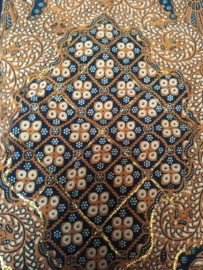 Fabric displays a symmetrical design with a quilted diamond pattern over a blue background. The diamonds contain orange quatrefoils surrounded by small white dots. Gold-colored thread accents the borders of the diamond shapes. The surrounding areas are a detailed gold and white pattern. Overall, it has the appearance of traditional batik textile art.