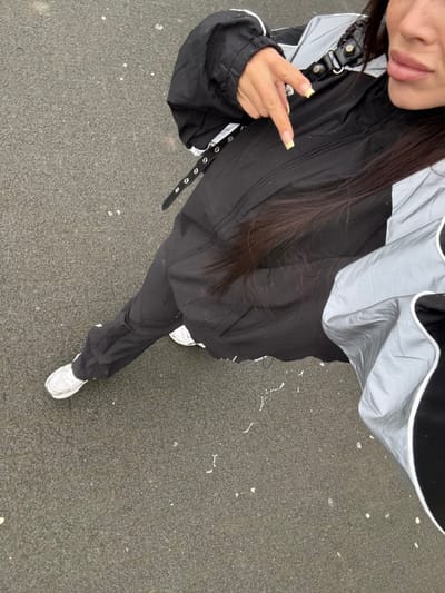 The image displays a partial view of an individual standing outdoors on a dark grey, textured ground surface in Sint-Niklaas, Belgium. The viewpoint is from above, looking downwards. The subject, a person with long, dark hair, is wearing a black jacket with reflective silver-grey panels and white piping, black trousers, and white sneakers with light grey elements. A black strap with multiple silver eyelets is visible across the upper torso. The person's right hand is raised, with the index finger pointing downwards towards the ground. Fingernails are light yellow. The lower right portion of the subject's face, including the jawline and lips, is visible. The ground surface shows scattered small white specks.
