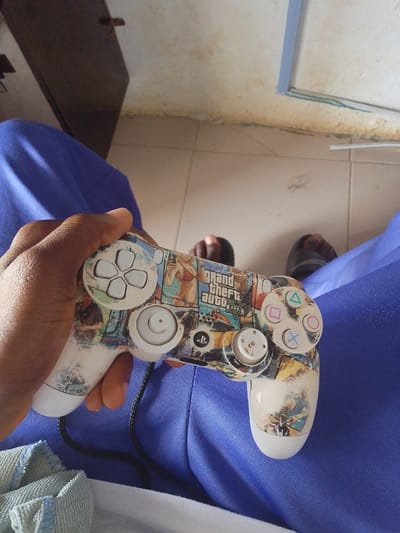 The image shows a person holding a video game controller that is decorated with images from the game Grand Theft Auto. The controller is white with a graphic skin featuring characters and artwork from the game. The person's hand, with dark skin, is holding the controller from the left side, with their thumb resting near the directional buttons.
The controller is placed on a surface of blue fabric, which appears to be clothing or upholstery. In the background, there are hints of a tiled floor and what looks like a doorway or opening leading to another room. Two pairs of feet, wearing sandals, are visible in the background, suggesting the presence of other people in the scene. One person is wearing brown sandals, and the other is wearing dark, possibly black, sandals.
The setting seems to be indoors, possibly a home or a common area. The lighting suggests it could be daytime, but there are no strong shadows to indicate a specific time. The overall atmosphere is casual, indicating someone is engaged in playing video games. The visible text on the controller clearly reads "Grand Theft Auto V." The scene is set in Abeere, Nigeria.
