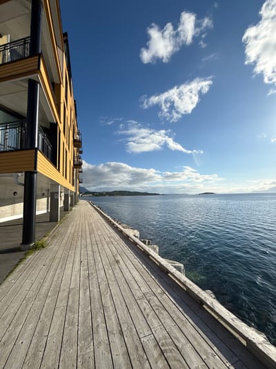 This image shows a boardwalk next to a body of water, with a building in the background. The boardwalk is made of wooden planks and runs parallel to the water. The building is modern, with a yellow facade and balconies. The water is calm and blue, and there are mountains and islands in the distance. The sky is blue with some scattered clouds, and the sun is shining, casting shadows. The scene appears to be in the city of Harstad, Norway, given the architecture and the coastal landscape. There are no people visible in the image. The environment is serene and picturesque, depicting a waterfront scene in what looks like a peaceful urban or semi-urban setting.