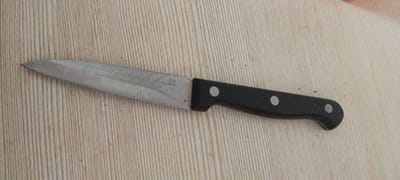 A stainless steel kitchen knife with a black handle is lying on a light-colored wooden surface. The knife is positioned horizontally, with the blade pointing to the left and the handle to the right. The blade appears slightly worn and has some faint scratches. There is text etched into the blade, but it is difficult to read clearly. The handle is black and has two round, silver rivets holding it together. The wooden surface has a subtle grain pattern. There are no people visible in the image. The lighting suggests an indoor setting, possibly a kitchen or dining area. The overall impression is of a common household object.
