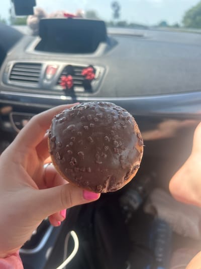 A close-up shot captures a hand holding a chocolate-covered donut inside a car. The donut, coated in dark chocolate and sprinkled with small, irregular brown pieces, is the primary subject. The hand, with bright pink nail polish, delicately grips the pastry. In the blurred background, the interior of the car is visible, featuring a dashboard with air vents and what appears to be a navigation screen. Outside the car, a hazy view of trees and a road suggests a location outside of a city, possibly on a highway or rural road. The lighting indicates it is daytime. The specific location in Tsalapitsa, Bulgaria, is not discernible from the visual information provided in the image.