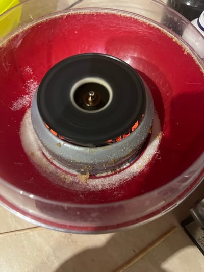 This close-up, overhead shot captures a red bowl with a clear plastic rim, containing a spinning black apparatus at its center. Granulated sugar is scattered around the base of the spinning apparatus and along the inner rim of the red bowl. The spinning mechanism appears to be part of a cotton candy machine, with heating elements glowing orange visible through its slats. A thin, wooden stick rests on the beige tiled surface next to the machine. The lighting suggests an indoor setting. No people are visible in the frame. There is no visible text.