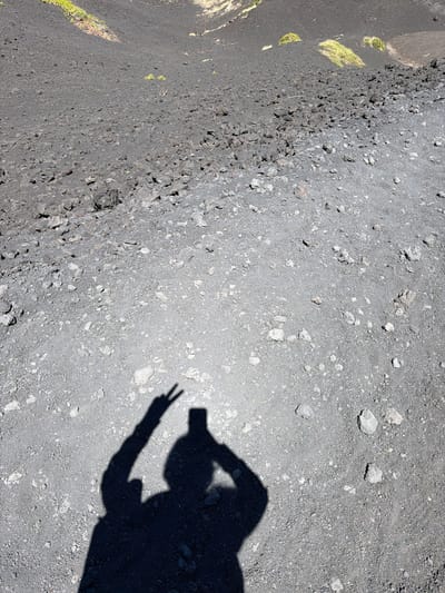 The image displays a shadow of a person on the rocky, volcanic terrain of Mount Etna, near Linguaglossa, Italy. The shadow, cast by the sunlight, is in the lower right quadrant of the image, showing a figure holding a smartphone and making a peace sign with their left hand. The terrain is composed of dark volcanic ash and scattered rocks of various sizes. In the background, patches of green vegetation are visible on the slope of the volcano. The lighting suggests it is a clear day with strong sunlight.