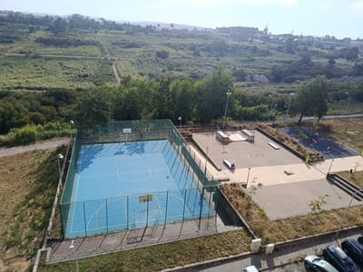 An aerial, high-angle shot captures a park area in Algueirão-Mem Martins, Portugal, on a clear day. In the foreground, a blue, fenced-in basketball court with white markings is visible. Adjacent to the basketball court is a skate park with various ramps and obstacles. A lone individual is seen in the skate park, sitting on a ledge. To the right of the skate park, a children's playground with red and black equipment is present. The park is surrounded by dry, grassy terrain and several trees. In the background, a hillside covered in vegetation and scattered buildings can be seen. Several parked cars are visible on a paved surface at the bottom right of the image.