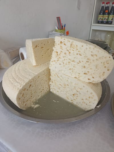 A large, round, off-white cheese with a patterned rind sits on a metal platter. The cheese has been cut, revealing a porous interior with numerous small holes. Two wedges of cheese are stacked on top of the round cheese, also displaying the porous texture. The platter contains a small amount of liquid, likely whey. In the background, a white wall is visible, with some plastic packaging to the left and a shelf with bottles and office supplies to the right. The setting appears to be a market or shop in Buynaksk, Russia.