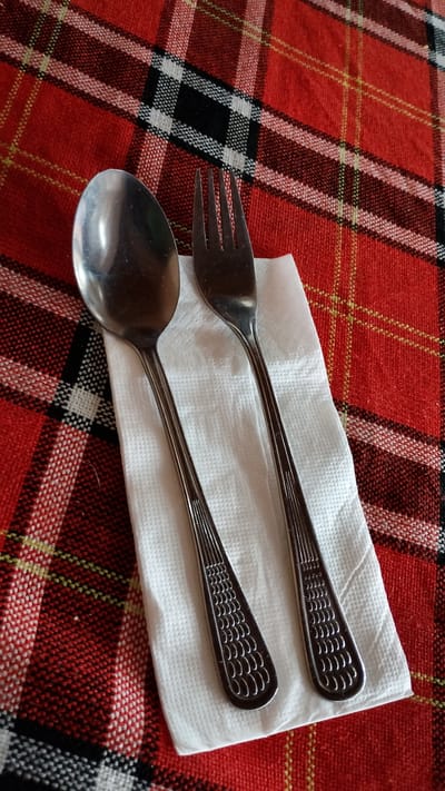A close-up, high-angle shot shows a fork and spoon resting on a white, textured napkin. The napkin is placed on a red and black plaid tablecloth. The cutlery is made of silver-colored metal and has a patterned design on the handles, featuring rows of small, curved lines resembling scales. The fork is to the right of the spoon, with its tines pointing upwards. The lighting is even, highlighting the reflective surfaces of the utensils and the texture of the napkin and tablecloth. There are no people present in the image. The scene appears to be set in a dining environment. The image was taken in Pevtsite, Bulgaria.