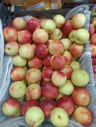 A high-angle, close-up shot shows a full frame of nectarines and peaches in a display crate at a market stall. The fruits are a mix of light yellow and red, with some showing a gradient of blush. The crate is lined with white, loosely woven fabric, and some of the fruit peeks out from under it. In the background, out of focus, there are shelves with packaged goods, including a bright yellow bag with Cyrillic writing. The overall impression is one of a fresh produce display in a market setting. This scene is located in the city of Gulbakhor, Uzbekistan.