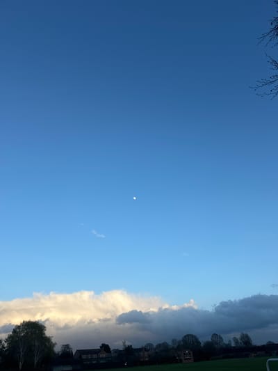 The image depicts a daytime sky above Easingwold, United Kingdom. A deep blue sky occupies the majority of the frame, with a small, pale moon visible. A vertical band of clouds, ranging from bright white to dark gray, fills the right side of the image. In the lower right corner, trees and rooftops of buildings are partially visible. A hint of green grass suggests a field or park area along the top right. Bare tree branches appear in the upper left corner. No people are visible.