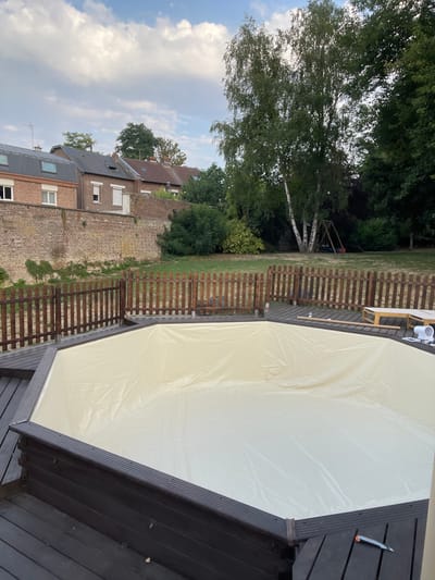 The image depicts an outdoor scene, likely a backyard, in the city of Chauny, France. In the foreground, a dark wooden deck surrounds an empty, hexagonal swimming pool lined with a pale yellow tarp. The pool structure appears to be made of dark wooden planks. Beyond the pool, a short, brown wooden fence encloses a grassy area. In the background, a brick wall with some overgrown ivy runs horizontally across the image. Behind the wall are residential buildings with various rooflines and windows, some with closed shutters. Further in the distance, a large birch tree with distinctive white bark stands prominently amongst other lush green trees. A children's swing set is visible behind the birch tree. The sky above is partially cloudy, with fluffy white clouds against a pale blue backdrop. The lighting suggests it is late afternoon or early evening. There are no people visible in the image.