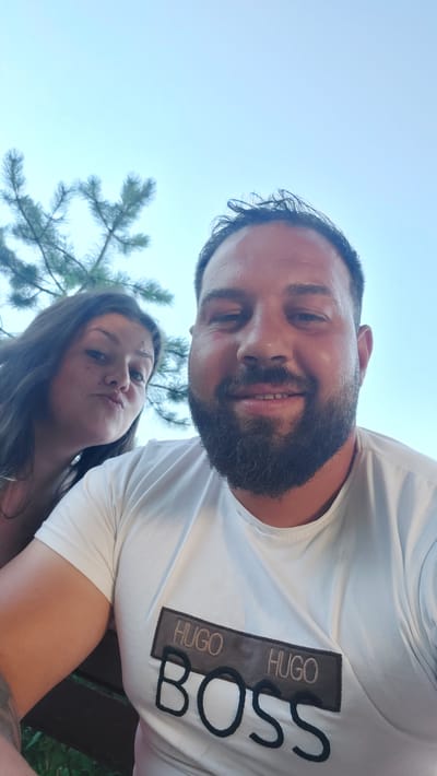 This media file is a selfie of a couple outdoors. The male subject is in the foreground, smiling and looking at the camera. He has a beard and is wearing a white t-shirt with a "BOSS HUGO BOSS" logo on it. Behind him, a female subject is leaning in and making a playful duck face. She has long, dark hair and is also wearing a white t-shirt. A tree branch is visible in the upper right corner of the image, suggesting a natural setting. The sky is a clear blue, indicating daytime. The overall mood of the image is happy and relaxed. The scene appears to be set in Suvorovo, Bulgaria.