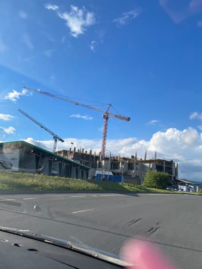 The image shows a construction site in Bittsa, Russia. The scene features unfinished buildings with multiple cranes extending into a bright blue sky with scattered clouds. There's a roadside visible, and some greenery near the construction site. The photograph was taken from inside a vehicle, as part of the car's interior is visible, including a portion of the dashboard and a pink reflection on the right.