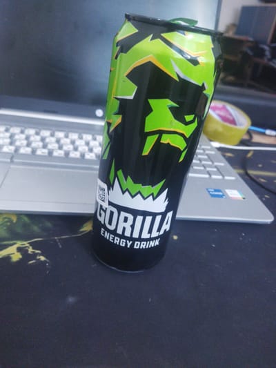 A tall, black can of "Gorilla Energy Drink" with a green and yellow jagged design is positioned on a dark mousepad in the foreground. The can is slightly tilted. In the background, a silver laptop is visible, with its keyboard exposed. A dark monitor is behind the laptop, and a blurred yellow object, possibly tape, is to the right of the can. The image is shot from a slightly elevated perspective. There is no indication of Zafar, Uzbekistan, in the image itself.