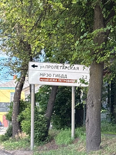 A street sign in Russian is visible in the center of the image, partially obscured by trees. The sign has white text on a white background, with some sections highlighted.

The top line of text reads "ул. ПРОЛЕТАРСКАЯ" (ul. Proletarskaya), which translates to "Proletarskaya Street." Below this, there is a black arrow pointing to the left.

The second line reads "МРЭО ГИБДД" (MREO GIBDD), which is an abbreviation for a traffic police department. To the right of this line, the number "2.2" is visible.

The third line has a brown rectangular background with white text that reads "МУЗЕЙ «ЕЖА ПЕТРОВИЧА»" (Muzey "Ezha Petrovicha"), translating to "Museum 'Ezha Petrovicha'."

The sign is attached to a metal pole and is surrounded by green foliage from trees, suggesting an outdoor setting likely during daytime, given the lighting. The image is taken from a slightly tilted angle. The background shows glimpses of buildings, one with yellow paint. The overall impression is of a typical street scene in a populated area, likely a town or city in Russia, given the language on the sign.