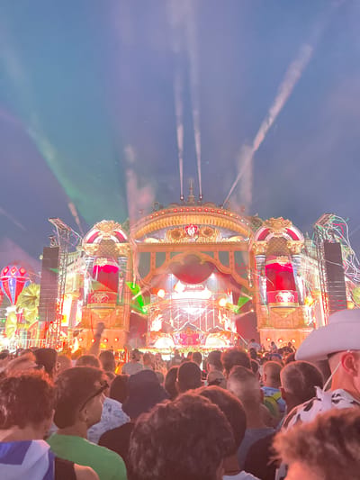 The image depicts a nighttime outdoor music festival scene, specifically at the Tomorrowland festival in Rumst, Belgium. A large, elaborate stage with intricate golden and red decorations dominates the background. The stage features multiple levels, spotlights, and what appears to be a red curtain with golden trim. Smoke or fog effects are visible, illuminated by green and white lights projected from the stage and into the sky, creating beams that extend upwards. In the foreground, a diverse crowd of festival-goers is visible from the back. People are dressed in casual clothing, with some wearing hats and some with their arms raised. The overall atmosphere is one of a vibrant, energetic event.