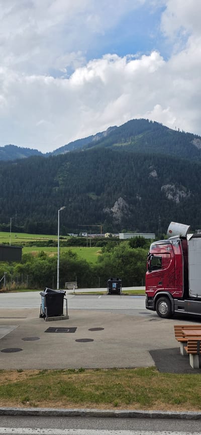 The image depicts a red semi-truck parked in what appears to be a service area or parking lot. The truck has a distinctive graphic on its side and a white cylindrical object protruding from its trailer. In the foreground, two black garbage bins are visible, one with its lid open. The ground is paved with concrete and features several circular manhole covers and a grate. A strip of dry grass with some green patches runs along the edge of the pavement.
Behind the service area, a line of green trees and bushes forms a natural barrier. Further in the background, a vast, densely forested mountain slopes upwards, its dark green foliage contrasting with the lighter green of the fields visible in the mid-ground. The sky is partially cloudy with patches of blue visible. In the distance, a crane and some buildings suggest a small town or industrial area nestled in the valley.
The scene is likely located in Kammern im Liesingtal, Austria, given the mountainous terrain and typical European architectural style suggested by the distant buildings. The overall impression is one of a rural or semi-rural setting, possibly near a highway rest stop or a logistics hub, with the imposing natural landscape dominating the background. There are no people visible in the frame.