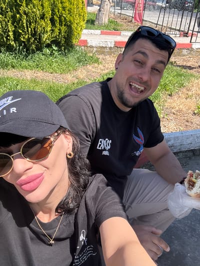 A man and a woman are posing for a selfie in an outdoor setting. The man is on the left, wearing a black t-shirt with an image of Bugs Bunny, sunglasses on his head, and he is holding a sandwich in a plastic bag. He is smiling broadly, exposing his teeth. The woman is on the right, wearing a black t-shirt, a black Nike cap, and sunglasses. She is wearing pink lipstick and a gold necklace. They are sitting on a grey concrete surface with red and white painted edging. In the background, there is grass and a bush on the left, and a grassy area on the right. There are cars and buildings visible in the distance, indicating an urban or suburban location. This is in Probuda, Bulgaria.